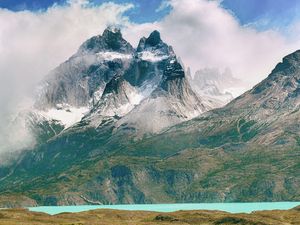 Jagged, snow-capped mountain peaks tower over a vibrant turquoise lake, with clouds partially obscuring the vast landscape. new Mapbox GL engine.