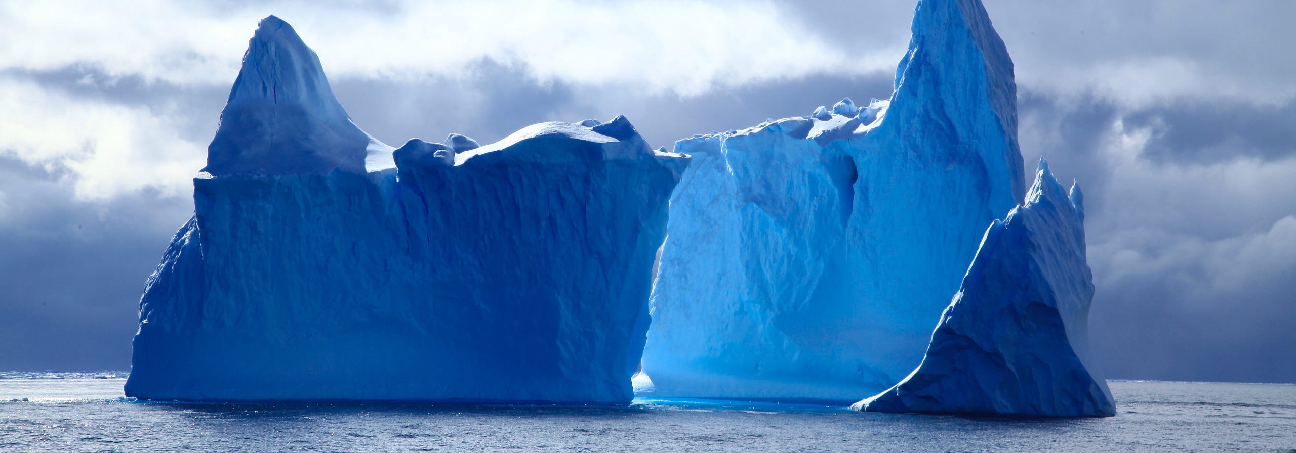 A panoramic photograph of massive, deep blue icebergs floating on the ocean under a dramatic and cloudy sky. MapHub's new Multi-Select tool.